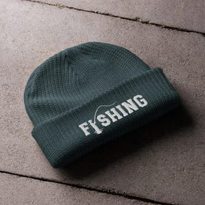 May include: A dark teal knit beanie with a folded cuff. The front of the cuff features white embroidered text that reads FISHING, with the letter I replaced by a graphic of a person holding a fishing rod and line extending over the letters.