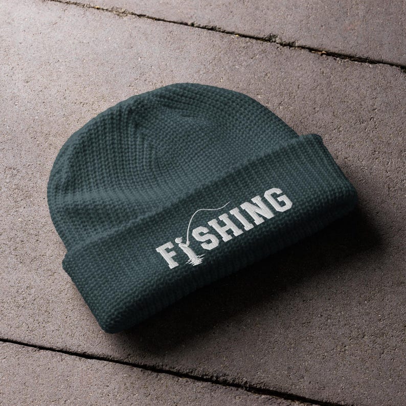 May include: A dark teal knit beanie with a folded cuff. The front of the cuff features white embroidered text that reads FISHING, with the letter I replaced by a graphic of a person holding a fishing rod and line extending over the letters.