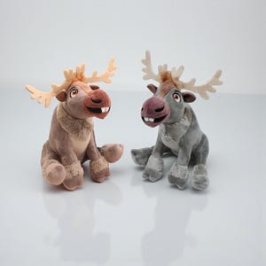 Pode incluir: Dois brinquedos de pelúcia em forma de alce. Um é marrom com chifres bege e um focinho marrom. O outro é cinza com chifres creme e um focinho roxo. Ambos têm uma expressão sorridente e estão sentados.