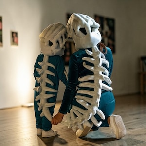 May include: Two people in matching dinosaur skeleton costumes. The costumes are teal with white bone detailing on the back and head. One person kneels, the other stands, holding hands. A fun, themed outfit.