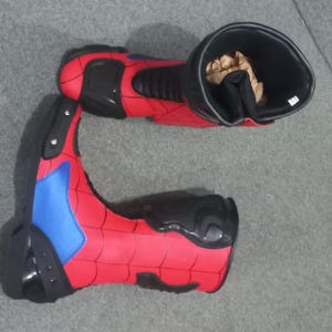 K&ouml;nnte beinhalten: Paar rote und schwarze Motorradstiefel mit blauem Akzent. Die Stiefel haben ein strukturiertes Design und Schutzelemente. Das Innere eines Stiefels ist sichtbar und enth&auml;lt braunes Verpackungsmaterial. Die Stiefel sind auf Sicherheit und Leistung ausgelegt.