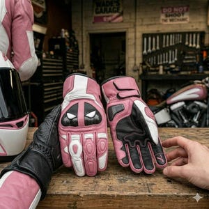 May include: A pair of motorcycle gloves featuring a pink, white, and black colour scheme. The gloves have a carbon fibre knuckle protector with a stylised white eye graphic, reinforced finger padding, and adjustable wrist straps for a secure fit.