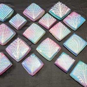 May include: An assortment of square-shaped cookies with iridescent pastel frosting in shades of blue, pink, and purple. Each cookie is decorated with a white, intricate design. The cookies are arranged on a dark surface.
