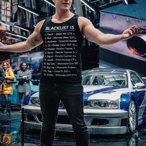 May include: A man wearing a black tank top with the text "BLACKLIST 15" stands in front of a blue and silver sports car. The tank top lists car models. The car has blue stripes and silver accents. The background is a car show.