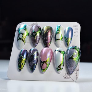 May include: A set of ten almond-shaped press-on nails displayed on a white card with size indicators XS, S, M, and L. The nails feature varied designs including white, pink, and dark glitter bases with iridescent green and black liquid-like patterns.