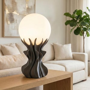 Puede incluir: Una lámpara de mesa moderna con una pantalla esférica de vidrio esmerilado. La base de la lámpara es un diseño oscuro y escultural con una forma orgánica y fluida. La lámpara está colocada sobre una mesa de madera en un salón.