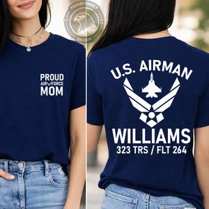 May include: Navy blue t-shirt with white text. The front reads "PROUD AIR FORCE MOM". The back displays "U.S. AIRMAN" above a graphic, with "WILLIAMS" and "323 TRS / FLT 264" below. The shirt has a crew neck.