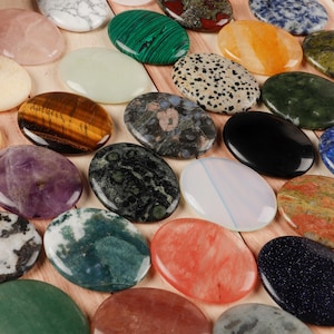 May include: A collection of polished, oval-shaped gemstones in various colors and patterns. The stones include rose quartz, tiger's eye, malachite, and black onyx. The gemstones are arranged on a light brown wooden surface.