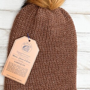 May include: A brown knitted beanie hat with a faux fur pom-pom. The hat has a tag that reads "Fenwood Fibre Company" and "Morris, Charlie & George". The tag also includes the text "A Labour of Love" and "Made in Newton, Ontario".