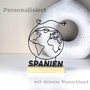 Könnte beinhalten: Schwarze Metallskulptur eines Globus mit Herz und Flugzeugdesign, montiert auf einem Holzsockel. Das Wort "SPANIEN" ist in schwarzen Buchstaben dargestellt. Der Text "Personalisiert" und "mit deinem Wunschland" sind ebenfalls sichtbar.