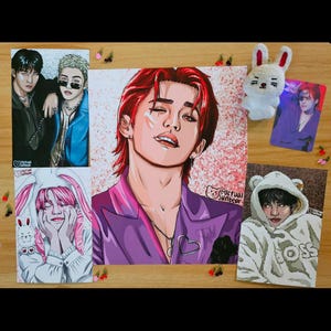 May include: A collection of fan art prints featuring stylized portraits of various individuals. The prints showcase vibrant colors and detailed illustrations, including a portrait with red hair and a purple jacket. A small plush bunny and a card are also visible.