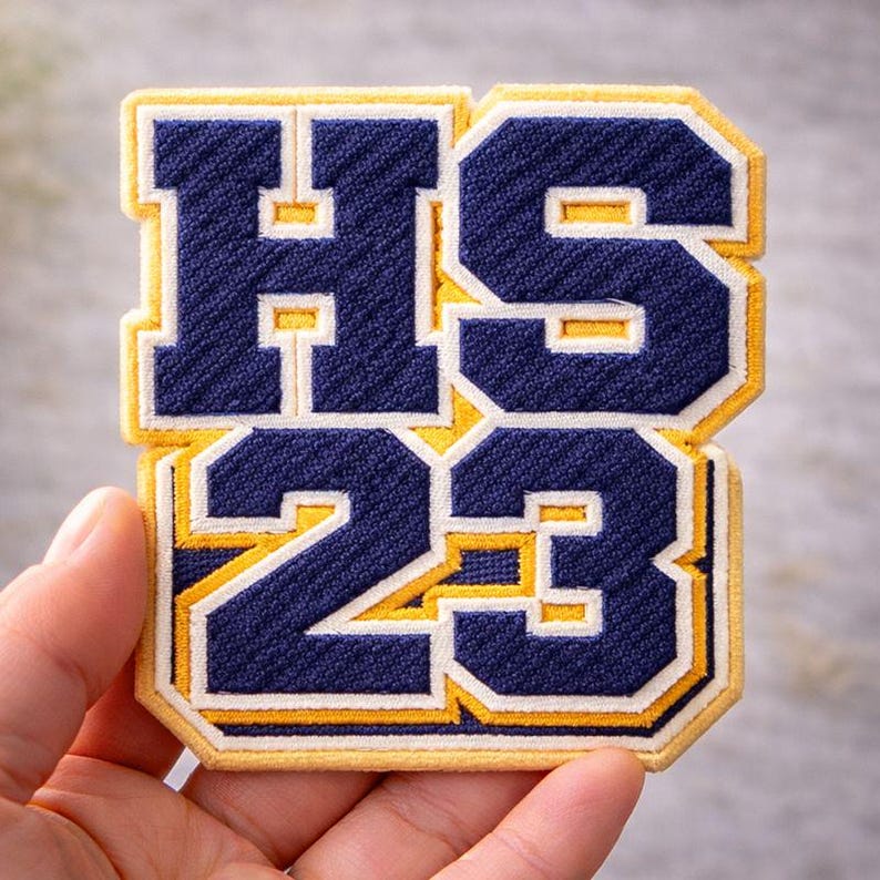 May include: An embroidered patch in navy blue and gold, featuring the letters "HS" above the number "23". The patch has a textured finish and a yellow border. The letters and numbers are in a bold, collegiate style.