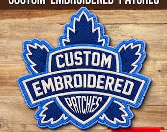 May include: A blue and white embroidered patch in the shape of a maple leaf. The patch features the words "CUSTOM EMBROIDERED PATCHES" in white lettering. The patch is set against a wooden background.