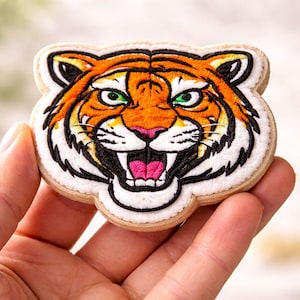 May include: Embroidered tiger head patch with orange, black, and white details. The tiger has green eyes, a pink nose, and an open mouth. The patch is on a light tan background.