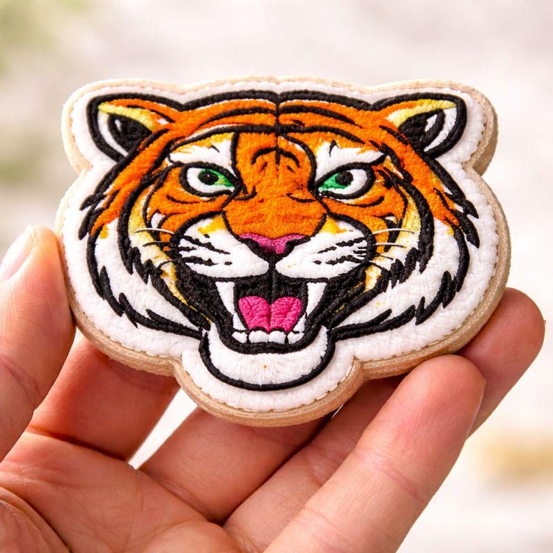 May include: Embroidered tiger head patch with orange, black, and white details. The tiger has green eyes, a pink nose, and an open mouth. The patch is on a light tan background.