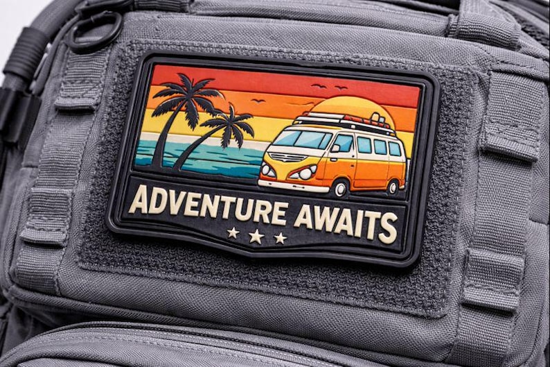 May include: A gray backpack with a rectangular patch featuring a colorful illustration of a vintage van on a beach with palm trees and the text "ADVENTURE AWAITS". The patch has a black border and three stars.