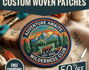 May include: A circular woven patch featuring an embroidered landscape with a brown bear, green pine trees, blue mountains, and an orange sunset. The text reads ADVENTURE AWAITS WILDERNESS CLUB EST. 2023 in a dark blue border with a stitched edge.