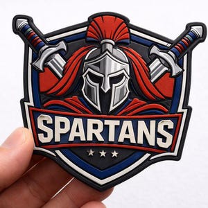 May include: A shield-shaped patch with a Spartan helmet and crossed swords design. The patch features the word "SPARTANS" in white letters, with red, blue, and black accents. Three stars are below the word.