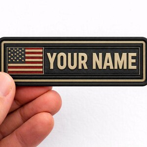 May include: A rectangular name tag with the text "YOUR NAME" in gold lettering. The tag features the American flag on the left side. The tag has a black border and is held by a hand.