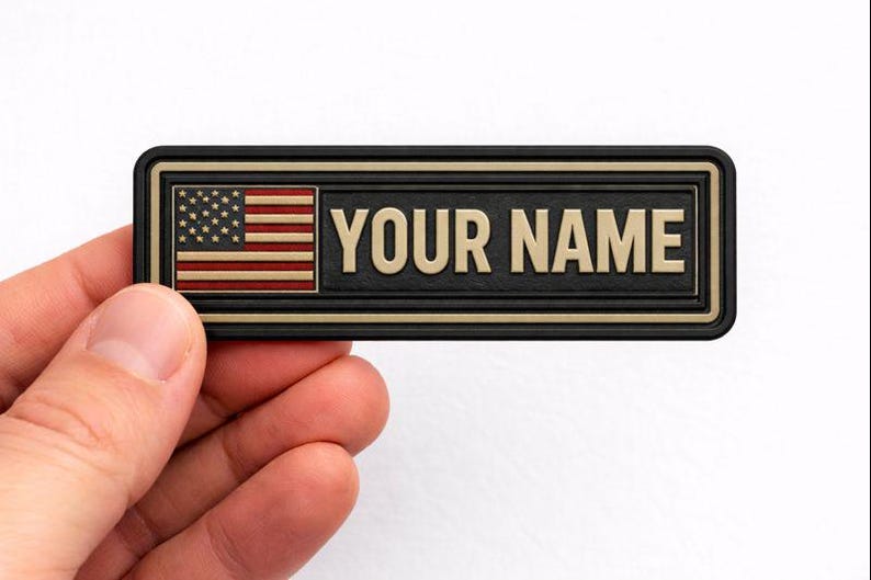 May include: A rectangular name tag with the text "YOUR NAME" in gold lettering. The tag features the American flag on the left side. The tag has a black border and is held by a hand.