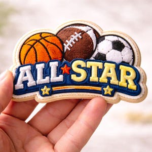 May include: Embroidered patch with a basketball, a rugby ball, and a football. The patch reads "ALL STAR" in white and yellow on a blue background, with star details. The patch is held in a hand.