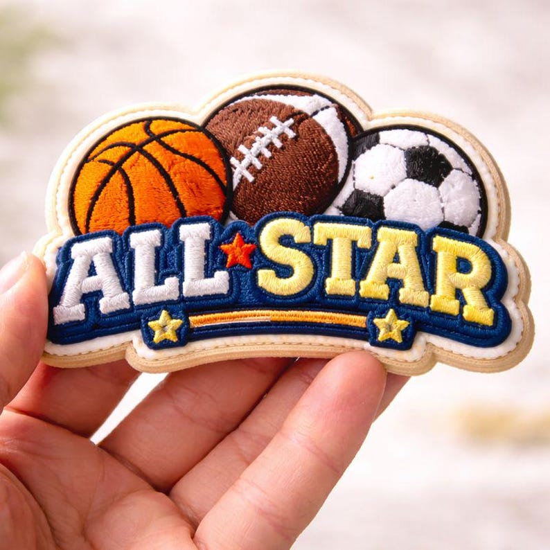 May include: Embroidered patch with a basketball, a rugby ball, and a football. The patch reads "ALL STAR" in white and yellow on a blue background, with star details. The patch is held in a hand.