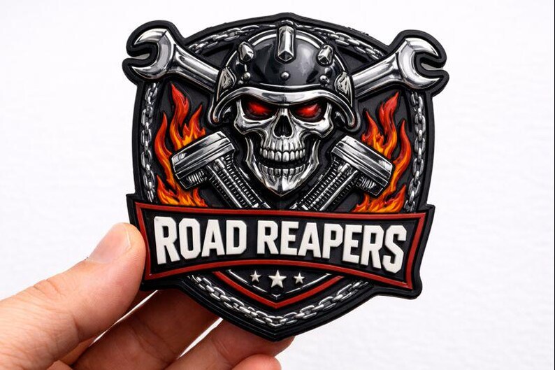 May include: A black and silver patch featuring a skull wearing a helmet with red eyes, crossed wrenches, and pistons. The patch has the words "ROAD REAPERS" in white on a red banner, with a chain border and flames.