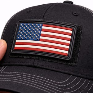 May include: Black baseball cap with a mesh back and a rectangular patch featuring the American flag. The flag design includes red and white stripes and a blue field with white stars. The cap is made of a durable fabric.