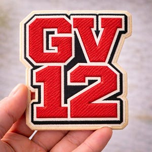 May include: A red and black embroidered patch featuring the letters "GV" and the numbers "12". The patch has a black border with a white outline and a beige background. The letters and numbers are in a bold, collegiate style.
