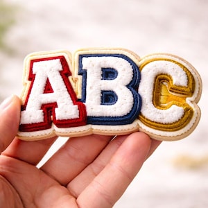 May include: A decorative patch spelling "ABC" in a varsity style. The letters are white with red, blue, and gold outlines, and have a textured, embroidered look. The patch is held in a hand.