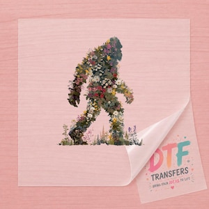 May include: A clear DTF transfer sheet featuring a silhouette of a Bigfoot creature composed entirely of colorful wildflowers and greenery. The bottom corner of the sheet is peeled back, revealing the text DTF TRANSFERS BRING YOUR IDEAS TO LIFE.