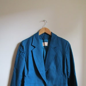 Puede incluir: Una chaqueta azul turquesa con un cierre de botón único y dos bolsillos. La chaqueta está colgada en una percha de madera.