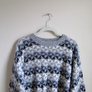Op de afbeelding: Een grijze, blauwe en zwarte geometrische patroon trui hangend aan een witte hanger. De trui heeft een herhalend driehoekontwerp in verschillende tinten blauw, grijs en zwart. De halslijn en manchetten zijn effen grijs.