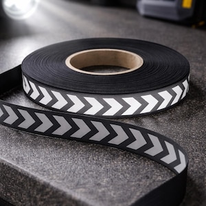 May include: A roll of black reflective tape with a repeating white chevron pattern. The tape is unrolled, showing the pattern. The background is a dark, textured surface. A flashlight is in the background.