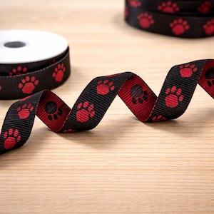 May include: Black ribbon with red paw prints, unspooled and in rolls. The ribbon is displayed on a light wood surface. The paw prints are evenly spaced along the ribbon, creating a repeating pattern. The ribbon is likely used for crafts or pet-related projects.