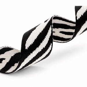 Op de afbeelding: Een zwart-wit gestreept zebra-lint, gekruld en vloeiend tegen een witte achtergrond. Het patroon van het lint is een gedurfd, herhalend ontwerp van zwarte strepen op een witte basis, waardoor een opvallend visueel contrast ontstaat. De stof lijkt geweven.