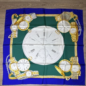 May include: A square silk scarf with a blue border, green center, and a white clock face design. Gold-colored pocket watches and wristwatches with Roman numerals and chains are arranged around the clock face. The text "Chopard Geneve" is visible.