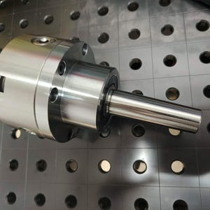 Puede incluir: Un mandril de torno de metal plateado con un cuerpo cilíndrico y un eje cónico. El mandril tiene múltiples orificios para pernos y un mecanismo de bloqueo. El objeto está sobre una superficie gris oscuro con agujeros espaciados.