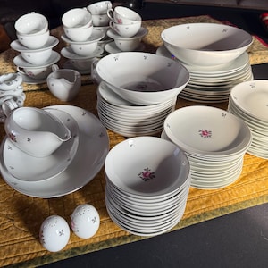 May include: A complete white porcelain dinnerware set with a delicate floral design. The set includes bowls, plates, cups, saucers, a gravy boat, and salt and pepper shakers. The set is arranged on a yellow quilted surface.