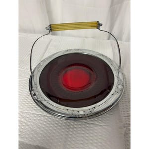Puede incluir: Una bandeja de servicio redonda con un inserto de vidrio rojo intenso en el centro, rodeado por un borde de metal cromado pulido con detalles en relieve. La bandeja incluye un asa de metal arqueada con una empuñadura de vidrio color ámbar.
