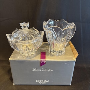 Può includere: Una zuccheriera in cristallo trasparente con coperchio e una lattiera abbinata della collezione Lotus di Gorham. Entrambi i pezzi presentano un design sfaccettato a forma di petalo. Sono esposti sopra una scatola grigia con il marchio Gorham.