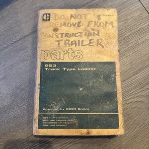 Puede incluir: Un manual rectangular para un cargador de cadenas 953 con una cubierta de color tostado y una sección rectangular de color verde oscuro. El texto dice: DO NOT REMOVE FROM CONSTRUCTION TRAILER parts, 953 Track Type Loader, Powered by 3204 Engine.