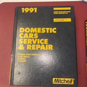 May include: A black book titled "1991 Domestic Cars Service & Repair, Volume 1" by Mitchell. The cover features yellow text and the Chrysler Motors Ford Motor Co. logo. The book covers engine performance, electrical, engine, and chassis.