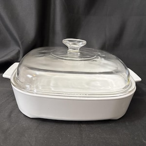 May include: A rectangular white ceramic baking dish with integrated side handles and a clear glass lid featuring a central knob handle. The dish has smooth, clean lines and a deep basin, designed for oven use and food preparation or serving.