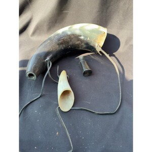 May include: A curved, dark-toned animal horn container with a polished surface, featuring a detachable threaded metal stopper and a separate, smaller, light-colored horn piece. A thin black leather cord is attached to the main horn body.
