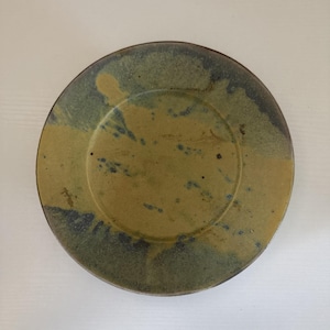 May include: A round ceramic plate with a speckled design. The plate features a yellow-brown center with dark blue and green edges. The plate has a slightly raised rim and a smooth surface.