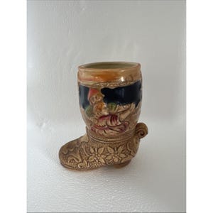 May include: A ceramic boot-shaped mug with a detailed floral design on the foot and a handle on the side. The upper portion features a painted scene with figures in various colors, set against a black and tan background.