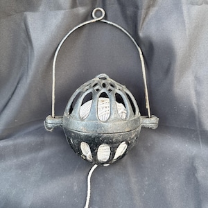 May include: A black metal yarn holder with a spherical cage design. The cage has decorative cutouts and a handle. Inside the cage is a ball of white yarn, with a strand extending out. The yarn holder is designed to hold and dispense yarn for knitting or crocheting.