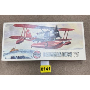 May include: A model kit box featuring a red and silver Grumman Duck seaplane. The box has the text "AIRFIX" and "GRUMMAN DUCK 72". A yellow sticker with the number "0141" is attached to the box.
