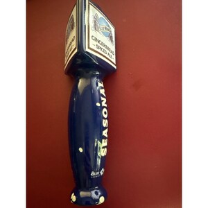 May include: A dark blue beer tap handle featuring a triangular top section with the text Blue Moon Gingerbread Spiced Ale. The cylindrical body of the handle is printed with the word Seasonal in white lettering and includes the Blue Moon logo.